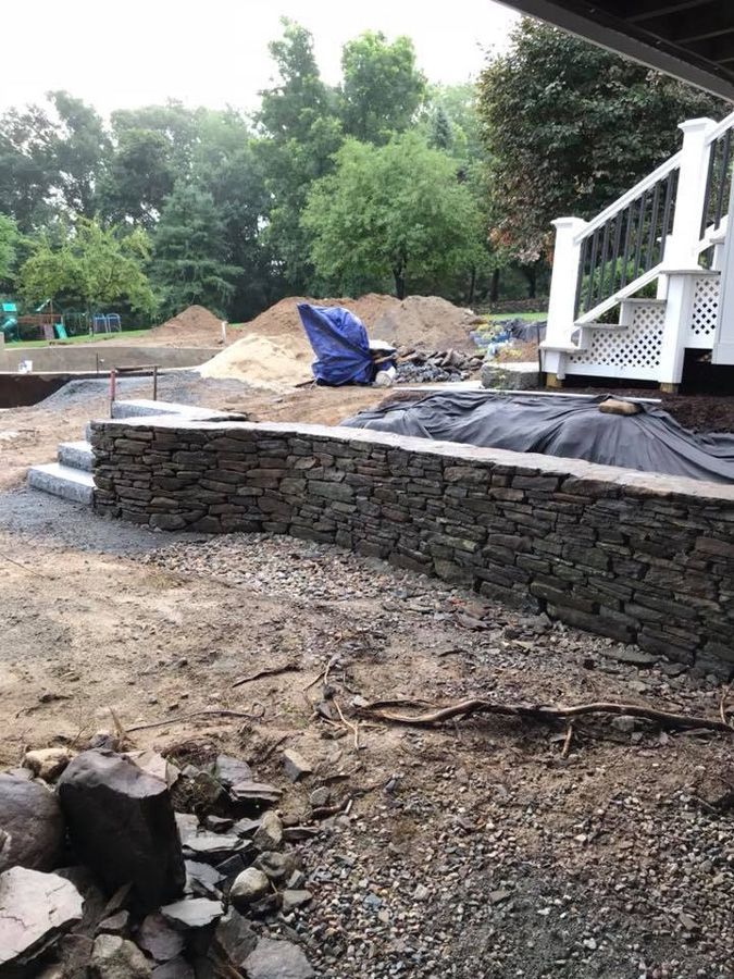 Stone retaining wall under construction near a white deck with steps, surrounded by dirt and gravel.
