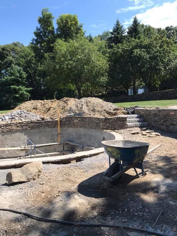 Pool construction site; stone walls, wheelbarrow, dirt, and trees against a bright blue sky.
