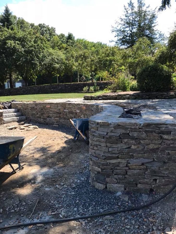 Stone retaining wall under construction in a garden setting with wheelbarrows and trees in the background.