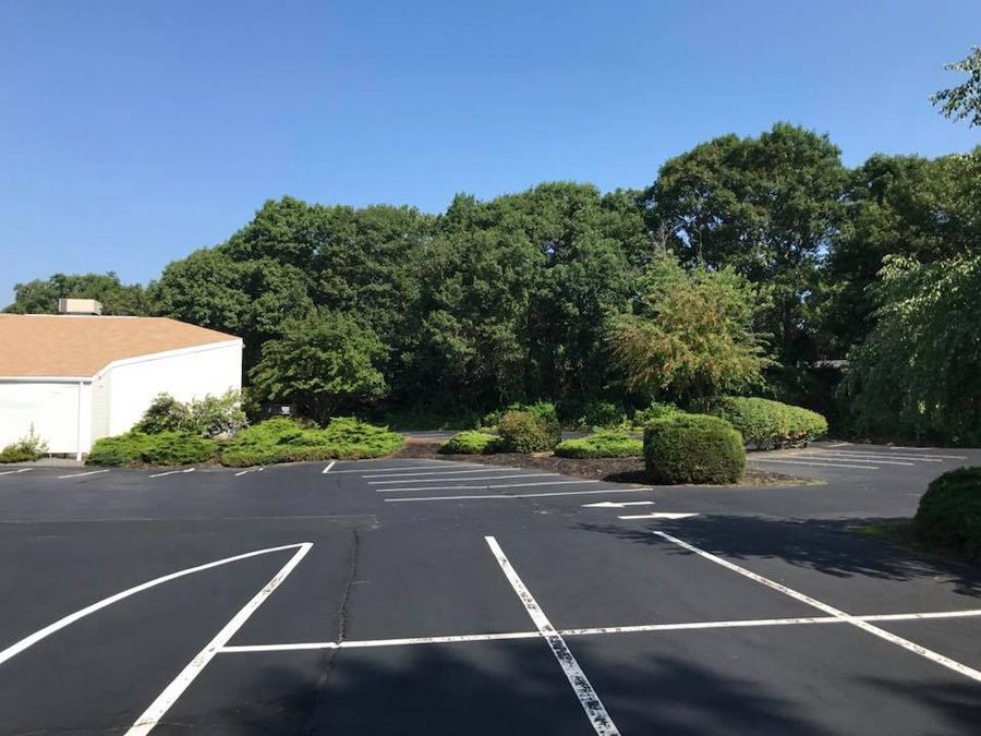 Empty asphalt parking lot with white lines, a building, and trees under a blue sky.