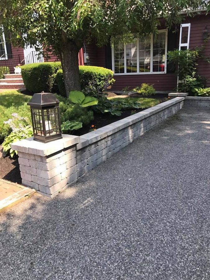 Gray brick retaining wall borders a garden next to a driveway, with a decorative lantern on the left.