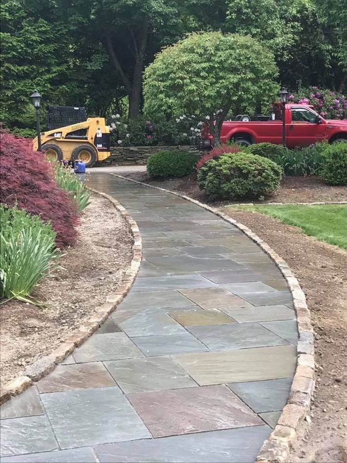 Stone path curves through a garden with a red truck, yellow loader, and flowering bushes.