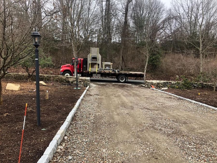 Red truck with a large gray structure on its flatbed, on a gravel driveway. Trees in the background.