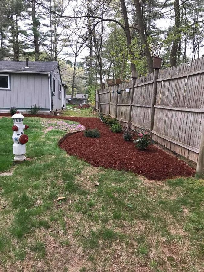 Red mulched flower bed next to a weathered wooden fence. Gray house and fire hydrant in the foreground.