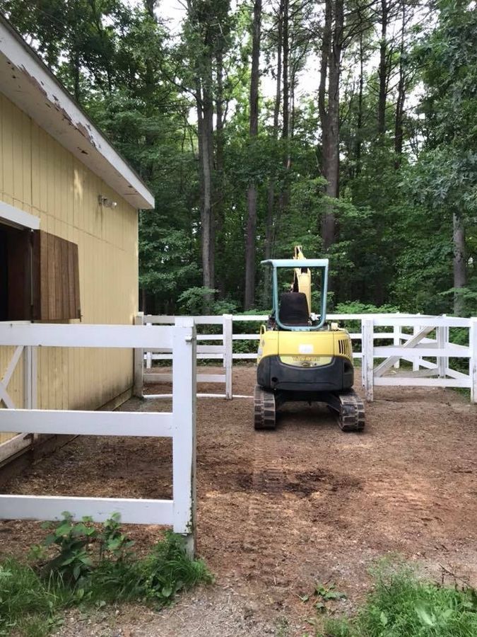 A yellow excavator inside a fenced area near a yellow building; trees in the background.