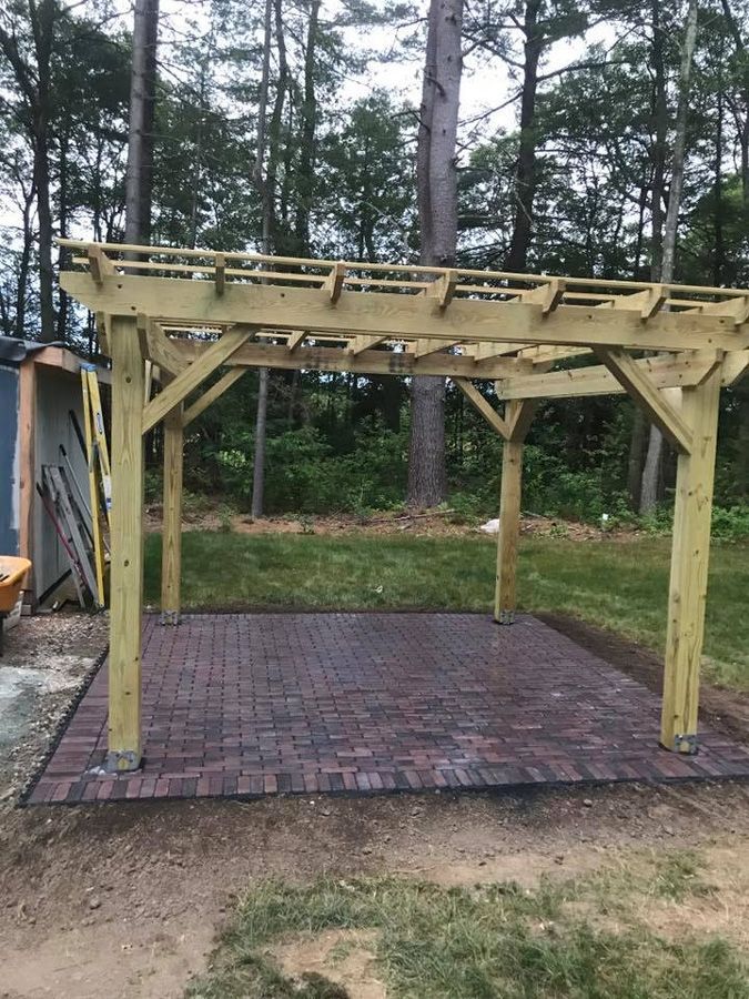 A wooden pergola over a brick patio in a backyard with trees.