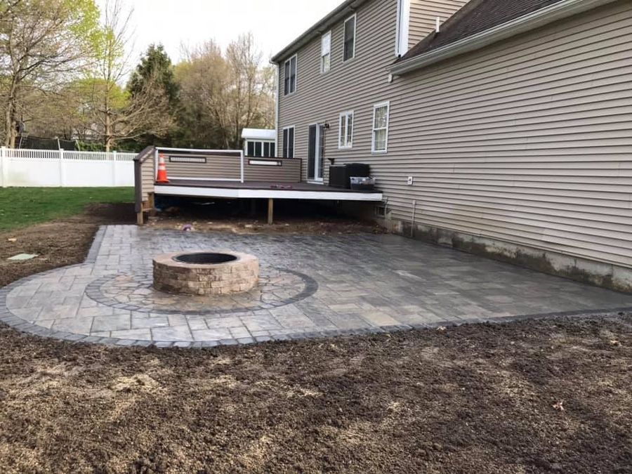 Backyard patio with fire pit, next to a house and deck, surrounded by grass.