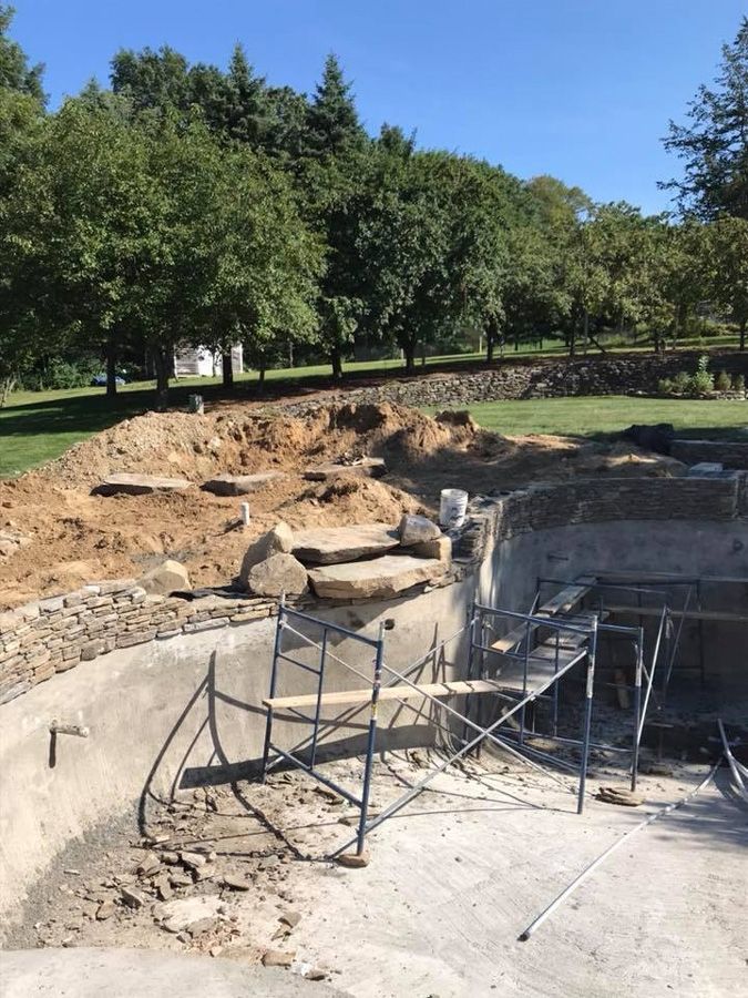 Pool construction site with scaffolding, stone steps, and exposed earth against a backdrop of trees.