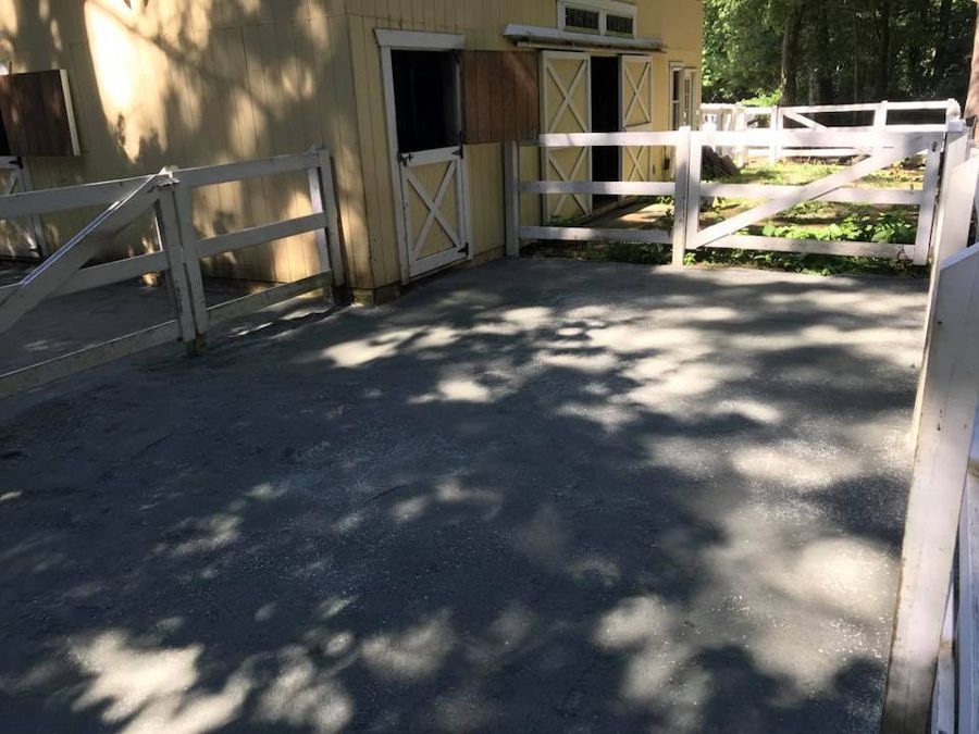 A light-yellow barn with open stalls and white fences. Sunlight casts shadows on the gravel ground.