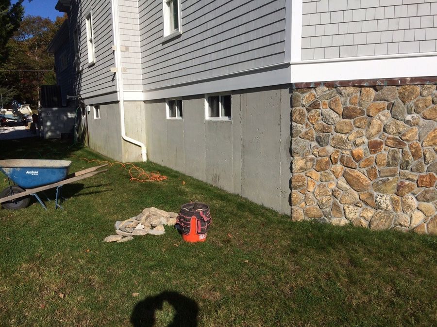 Side of a house with gray siding, stone foundation, and grass. A wheelbarrow, bucket, and debris are on the lawn.