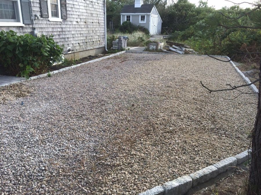 Gravel driveway edged with stone bordering a gray house and a small shed in a yard.