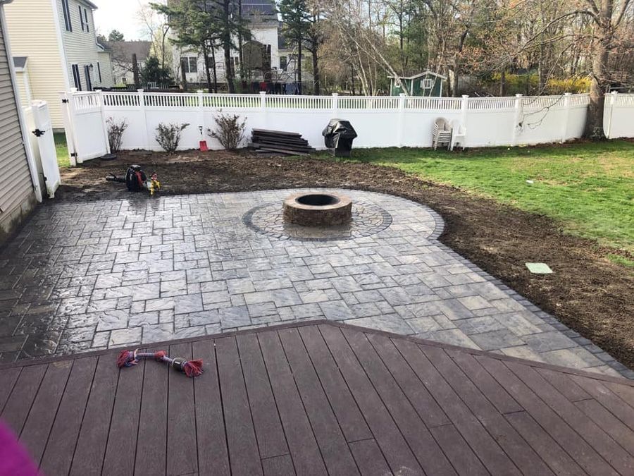 Backyard patio with stone pavers, fire pit, and composite deck. White fence and green grass surround.