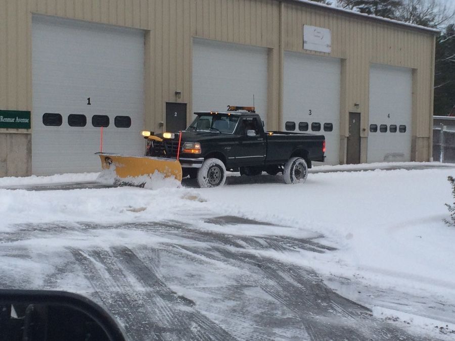 A dark pickup truck with a snowplow clearing snow in front of a building with garage doors.
