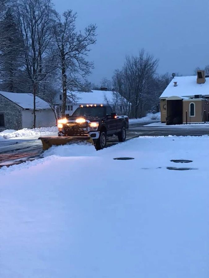 Snowplow truck clearing a snowy road; houses in the background under a cloudy sky.