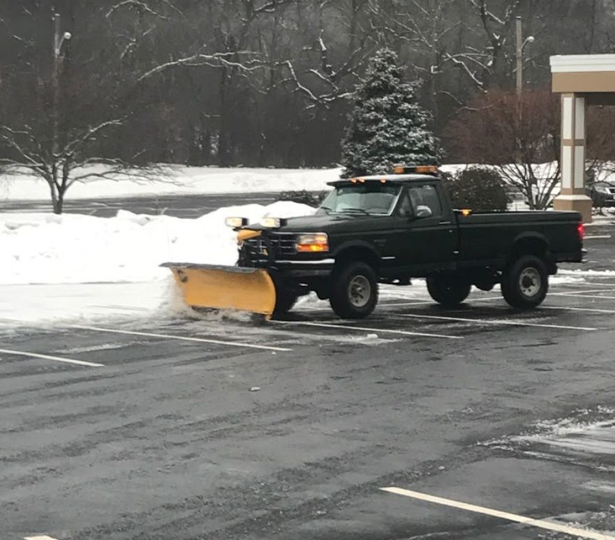 Green pickup truck with a snowplow clearing a parking lot on a snowy day.