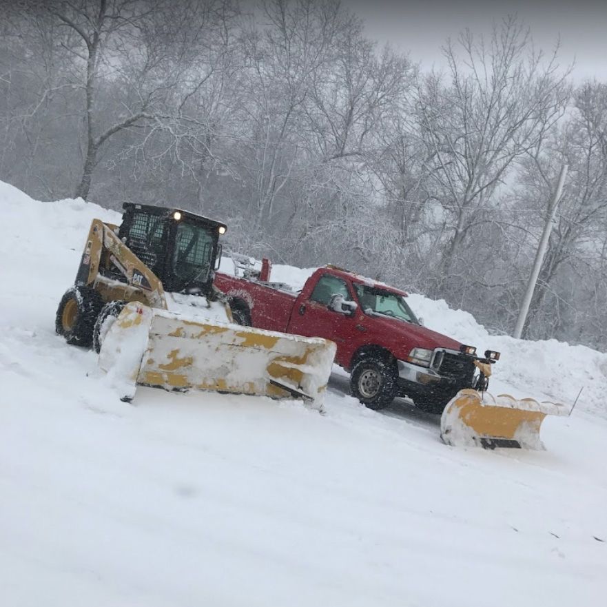 Snow-covered hillside with a red pickup truck and yellow skid steer both with snowplows clearing snow.