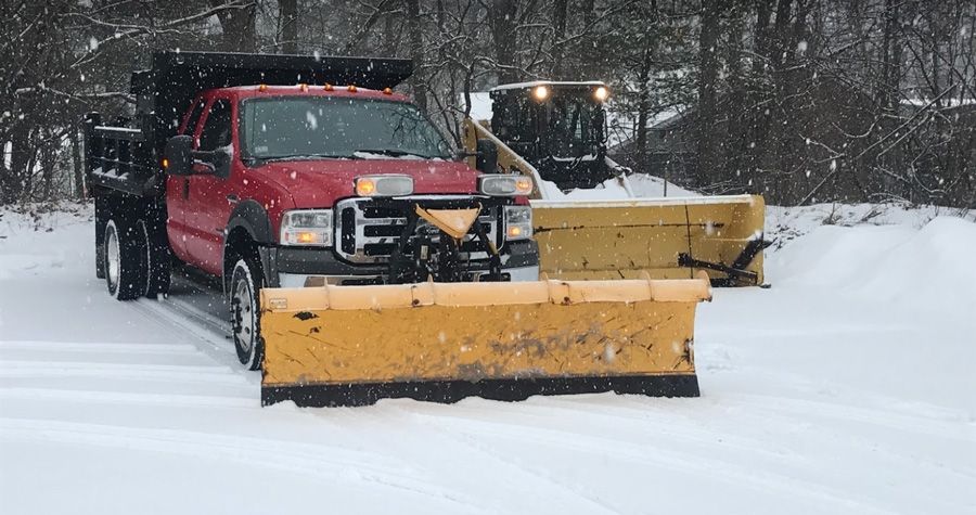 A red truck with a yellow snowplow clears snow during a snowfall; another truck is behind it.