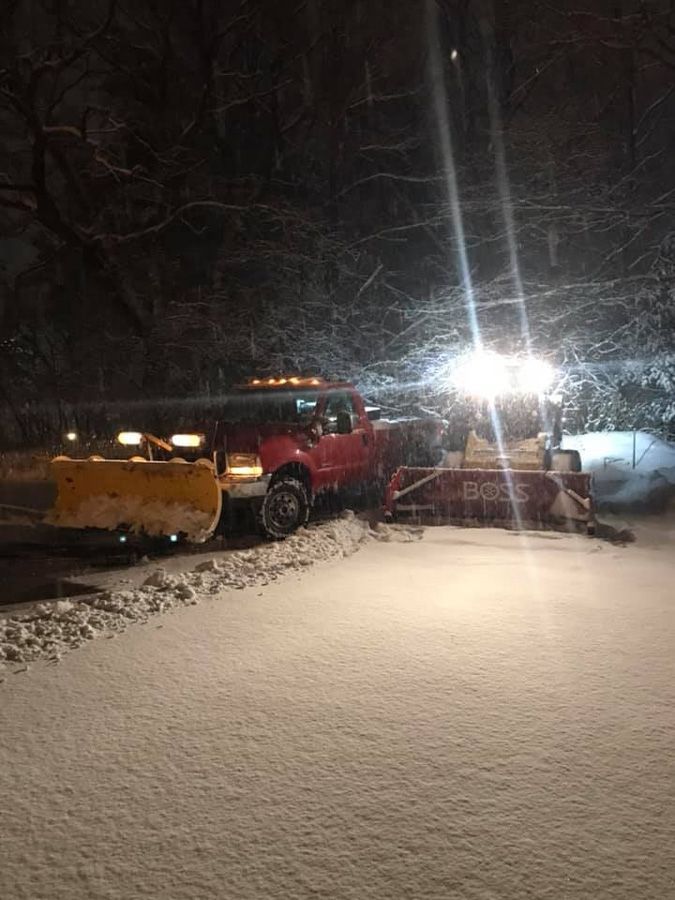 Red truck with snow plow clearing a snowy road at night.