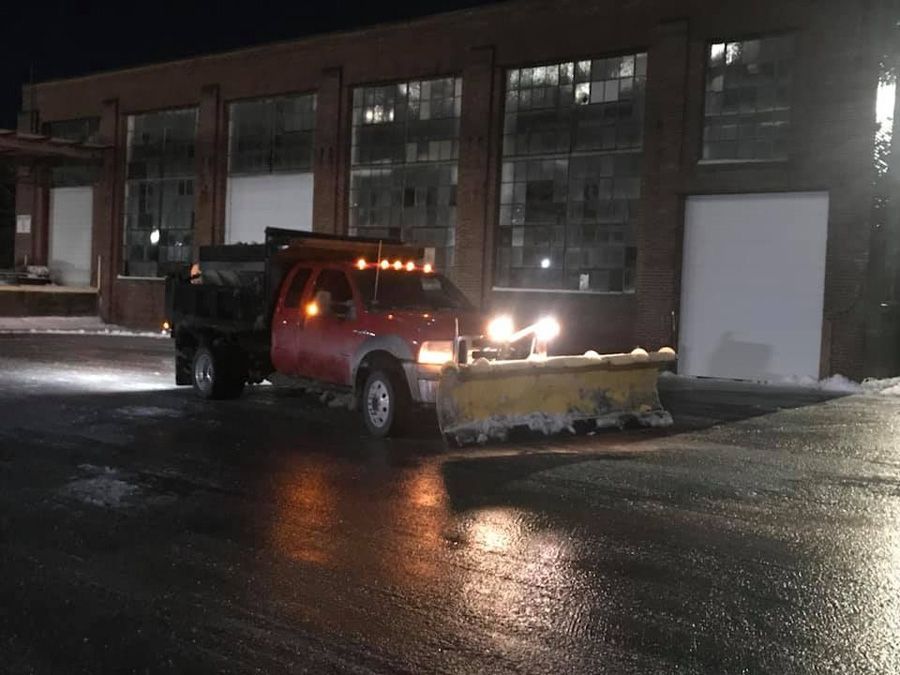 Red snowplow truck in front of a brick building at night, plowing a wet road.