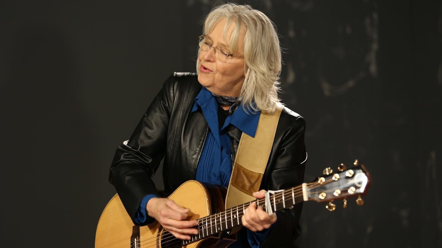 Musician playing acoustic guitar on stage, wearing a black jacket and blue shirt, against a dark background