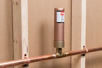 A copper water hammer arrestor installed on a horizontal copper pipe between two wooden wall studs.