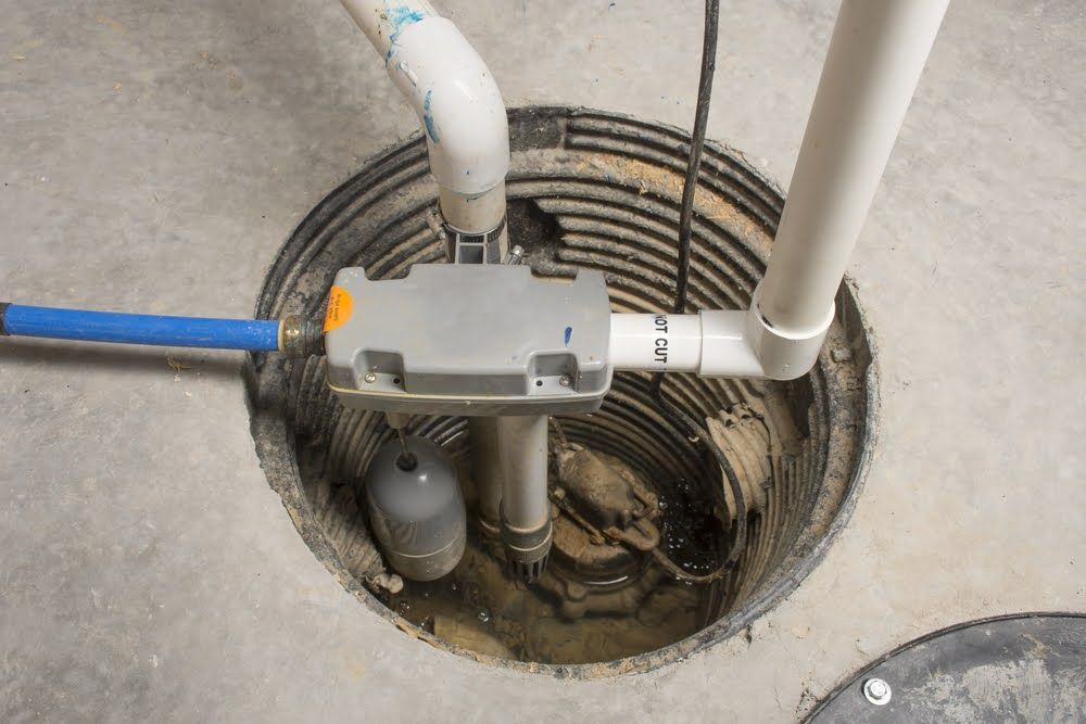 A sump pump system inside a concrete floor basin, with white PVC pipes, a grey control box, and a blue hose connection.