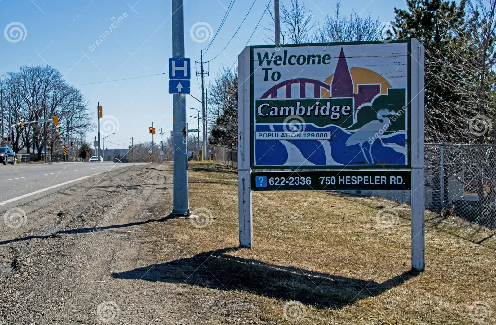 Sign welcoming people to Cambridge, Ontario, with a cityscape graphic, beside a road, blue sky.