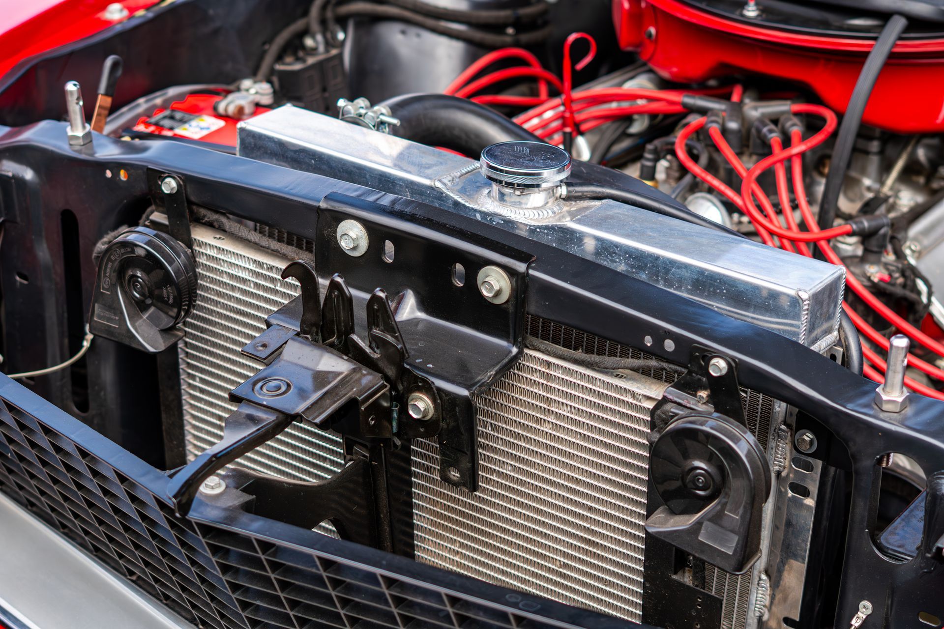 Close-up of a car engine bay with a black radiator, red wiring, and metal brackets.