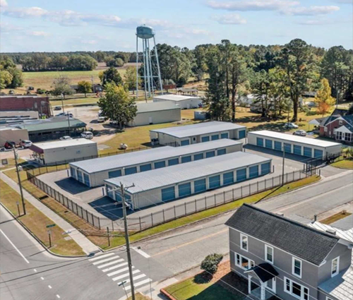 Aerial view of a self storage facility with low white buildings, blue windows, trees, and a water tower nearby