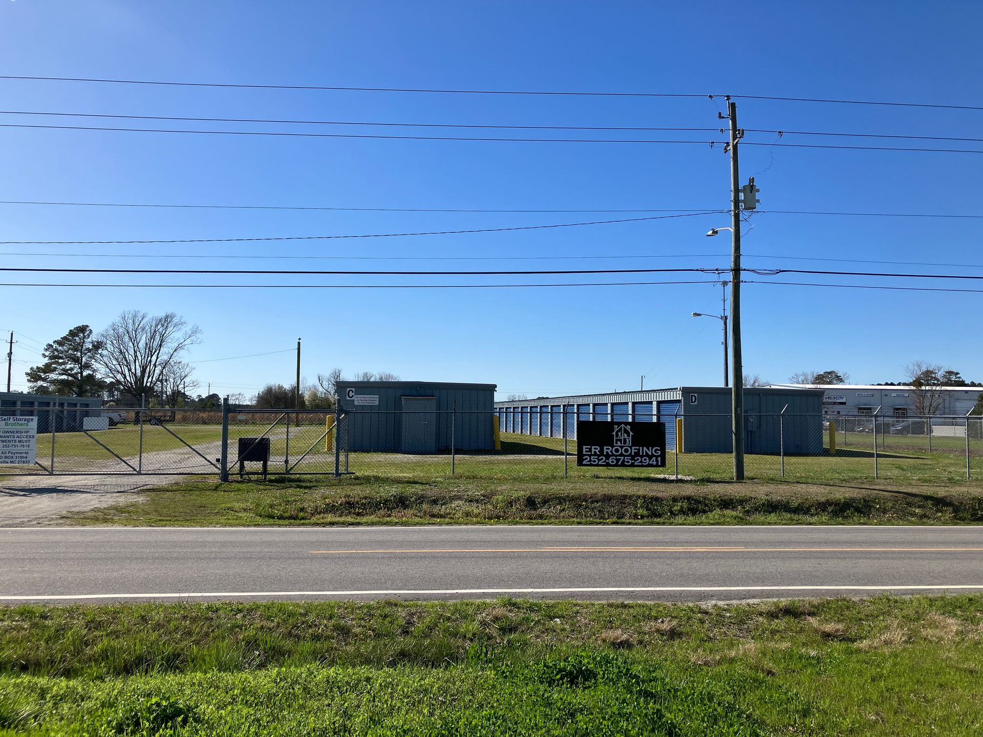 Self Storage with chain-link fence, blue sheds, utility pole, and parked sign under a clear sky