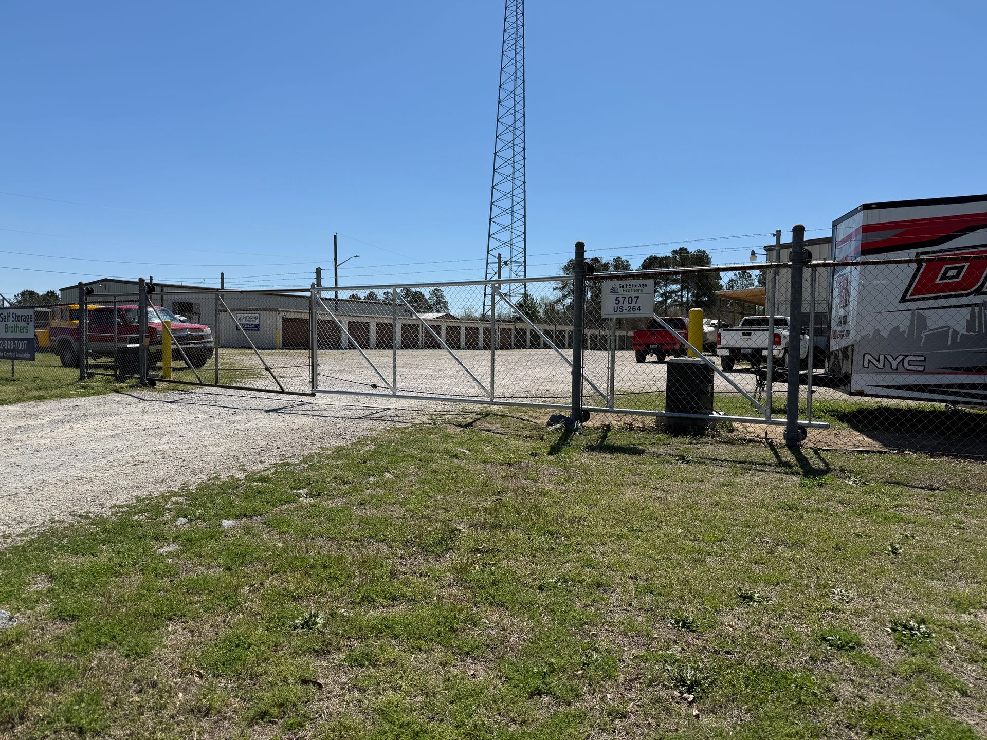 Closed chain-link gate at a gravel lot with utility pole and red-and-white barrier tape under a blue sky
