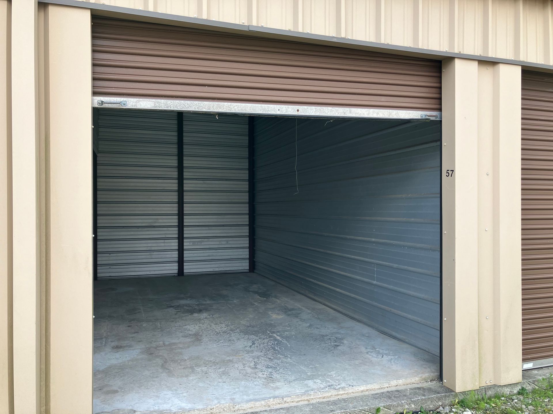 Open self storage unit with beige exterior, gray interior, and partially raised brown roll-up door