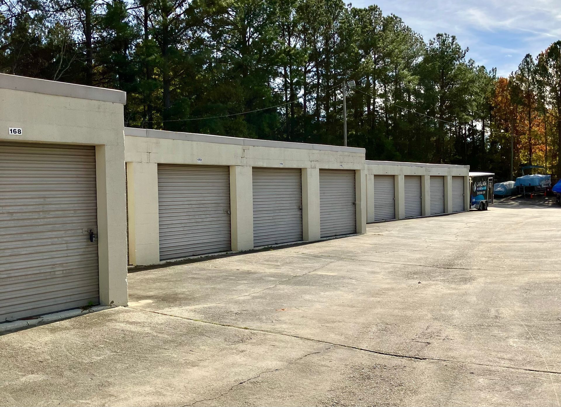 Row of beige storage unit doors with roll-up shutters along a gravel lot by trees
