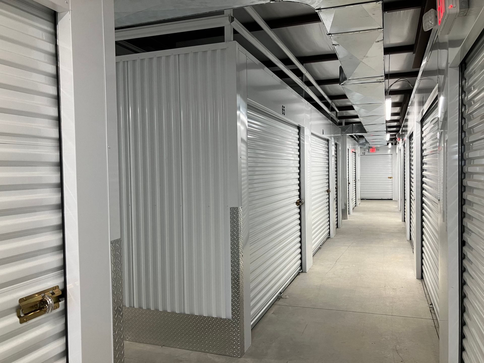 A row of white storage unit doors in a narrow indoor hallway with concrete floors and fluorescent lighting