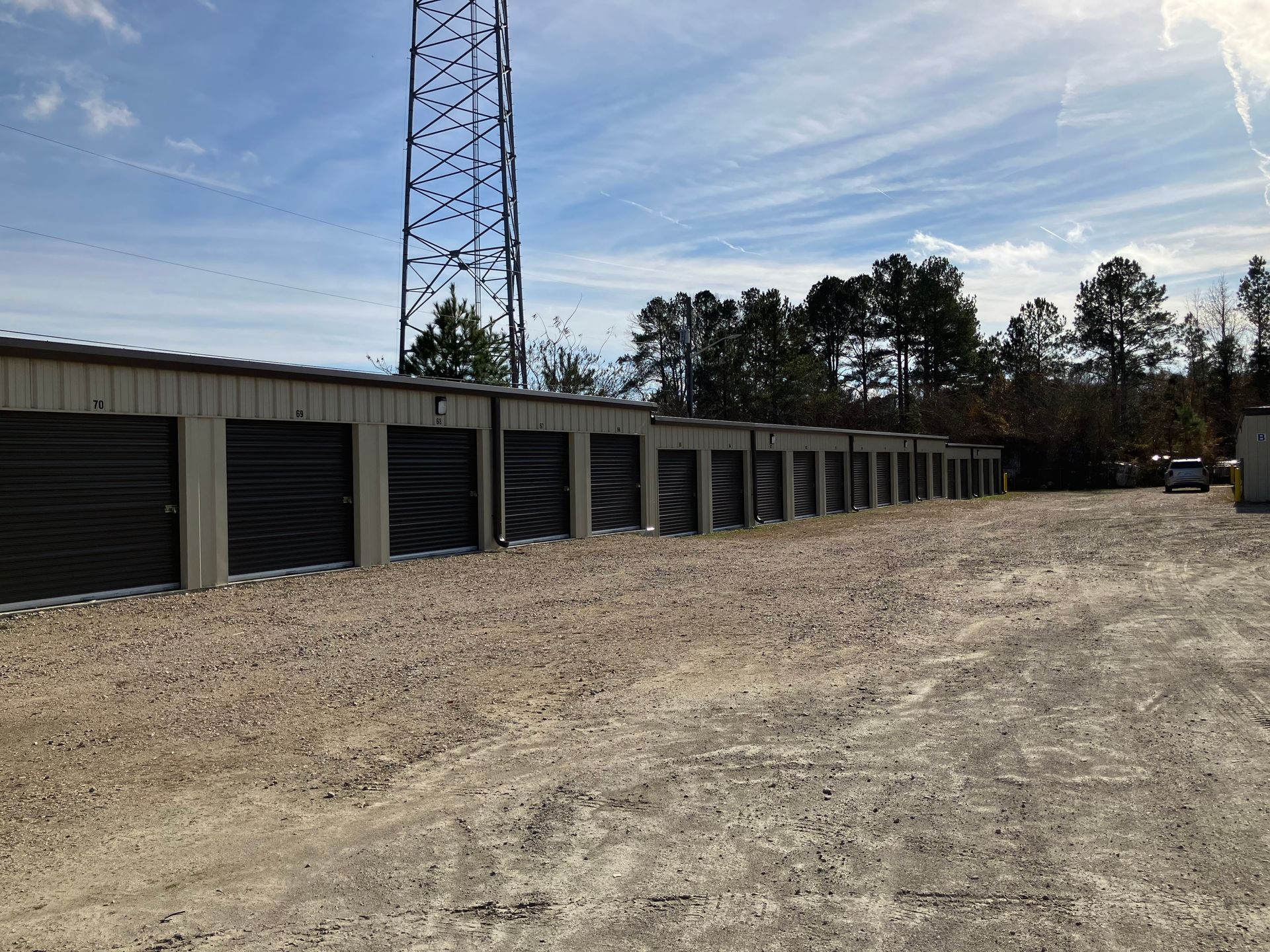 Row of storage units beside a dirt lot with a tall tower and trees under a blue sky