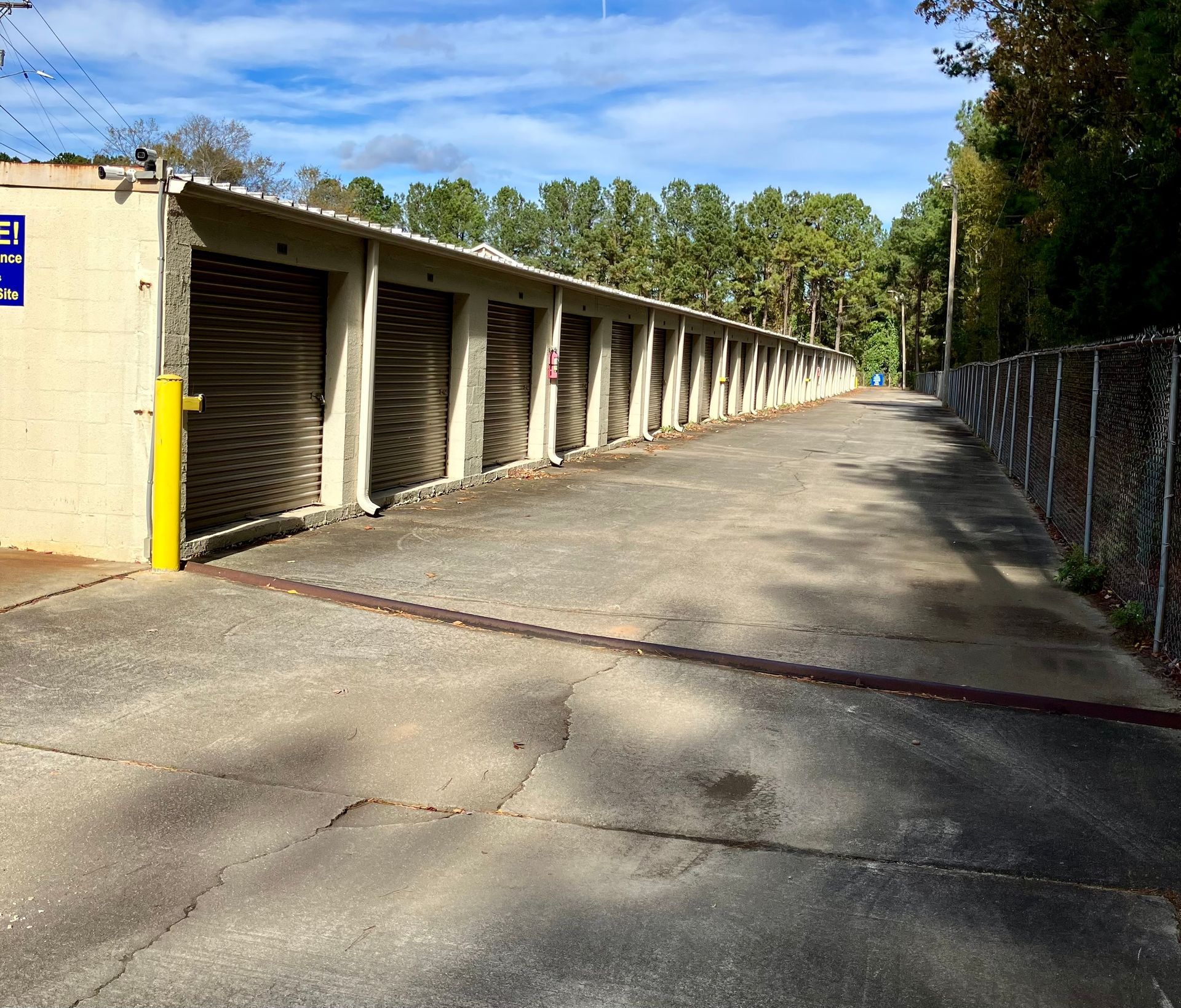 Row of beige storage unit doors along a paved drive under a blue sky, beside a chain-link fence and trees