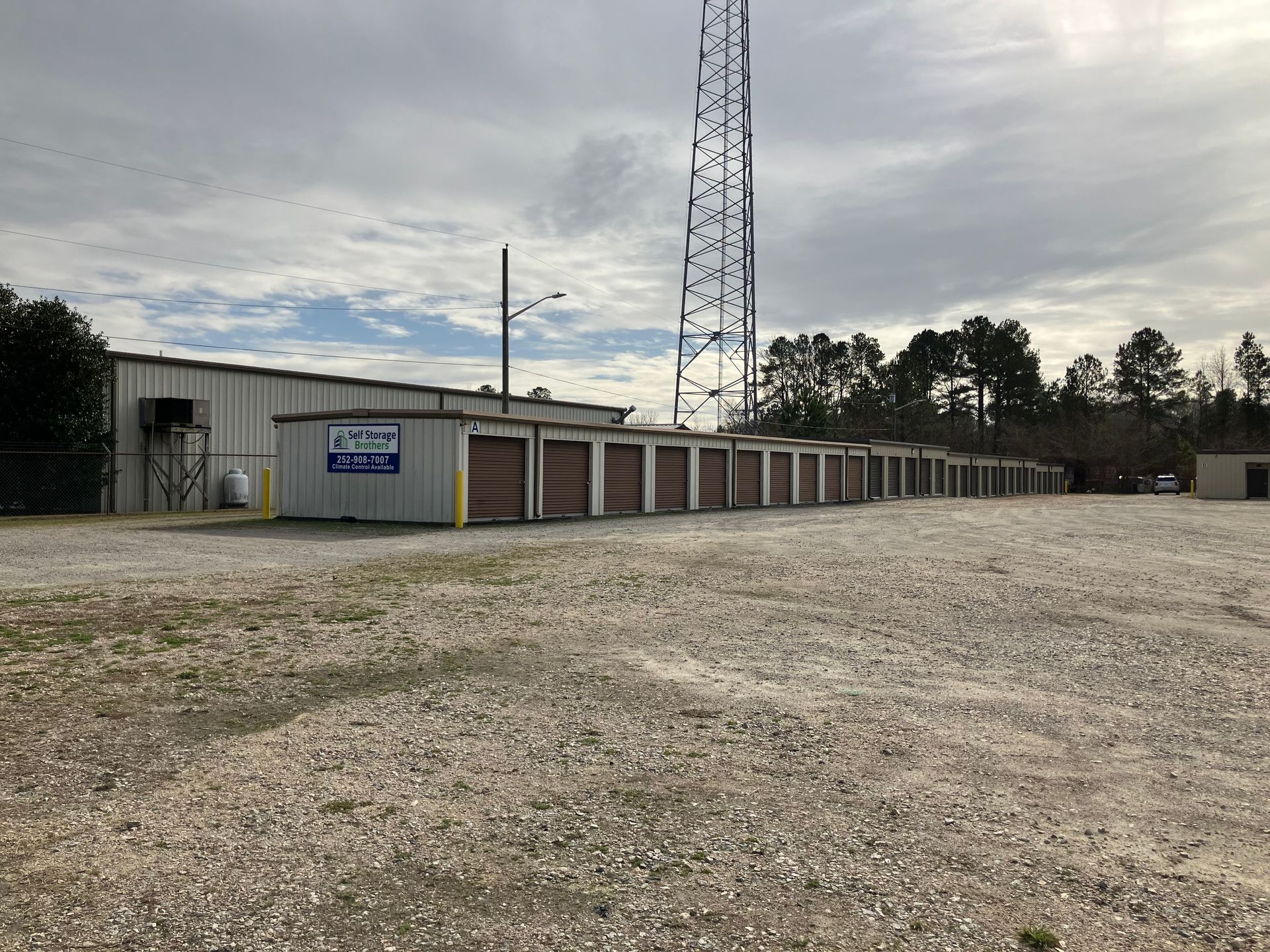 Gravel lot beside a row of storage units and a tall radio tower under a cloudy sky