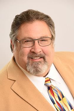 Smiling man in tan suit and colorful tie, wearing glasses, against a light background