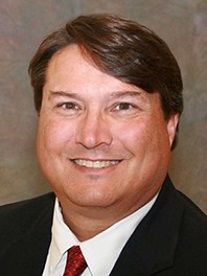 Smiling man in a black suit, white shirt, and red tie against a neutral background