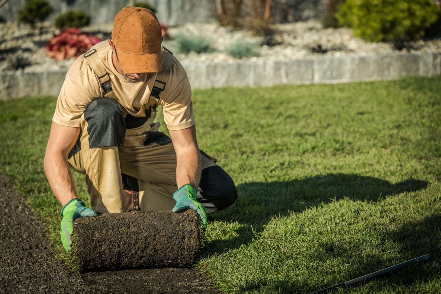 Person rolling out fresh sod on a lawn with green gloves and an orange cap.