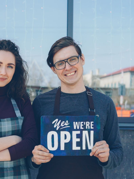 Two people standing outdoors holding a sign that says “We’re Open”