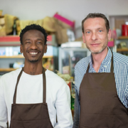 Two smiling shop workers in brown aprons stand behind a counter in a store.