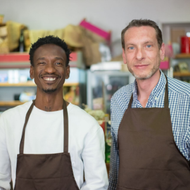 Two smiling shop workers in brown aprons stand behind a counter in a store.