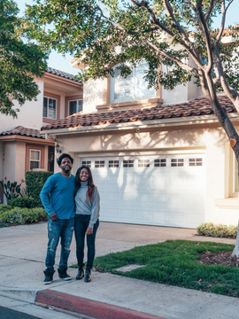 Two people standing on a suburban sidewalk in front of a white garage and beige house.