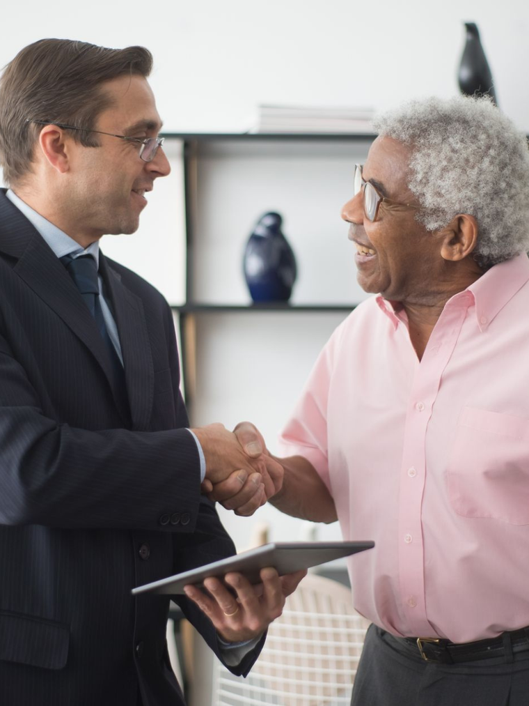Two professionals shaking hands in an office, one holding a tablet and the other in a pink shirt.