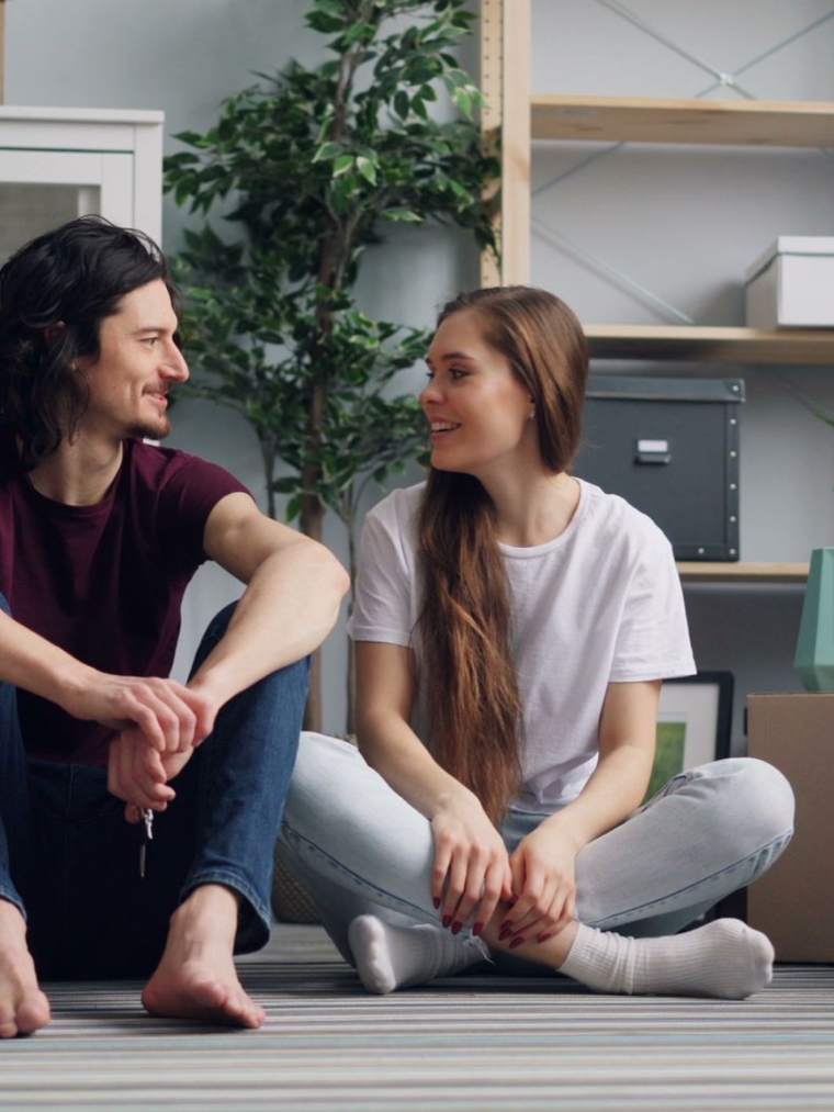Two people sit on a rug in a living room surrounded by moving boxes, smiling and talking.