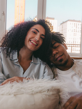 Two people smiling on a couch by a window with city buildings in the background