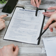 Hands reviewing a home insurance policy form on a clipboard with a pen on a table