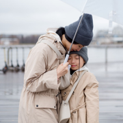 Two people in beige coats and gray beanies share an umbrella by the waterfront.