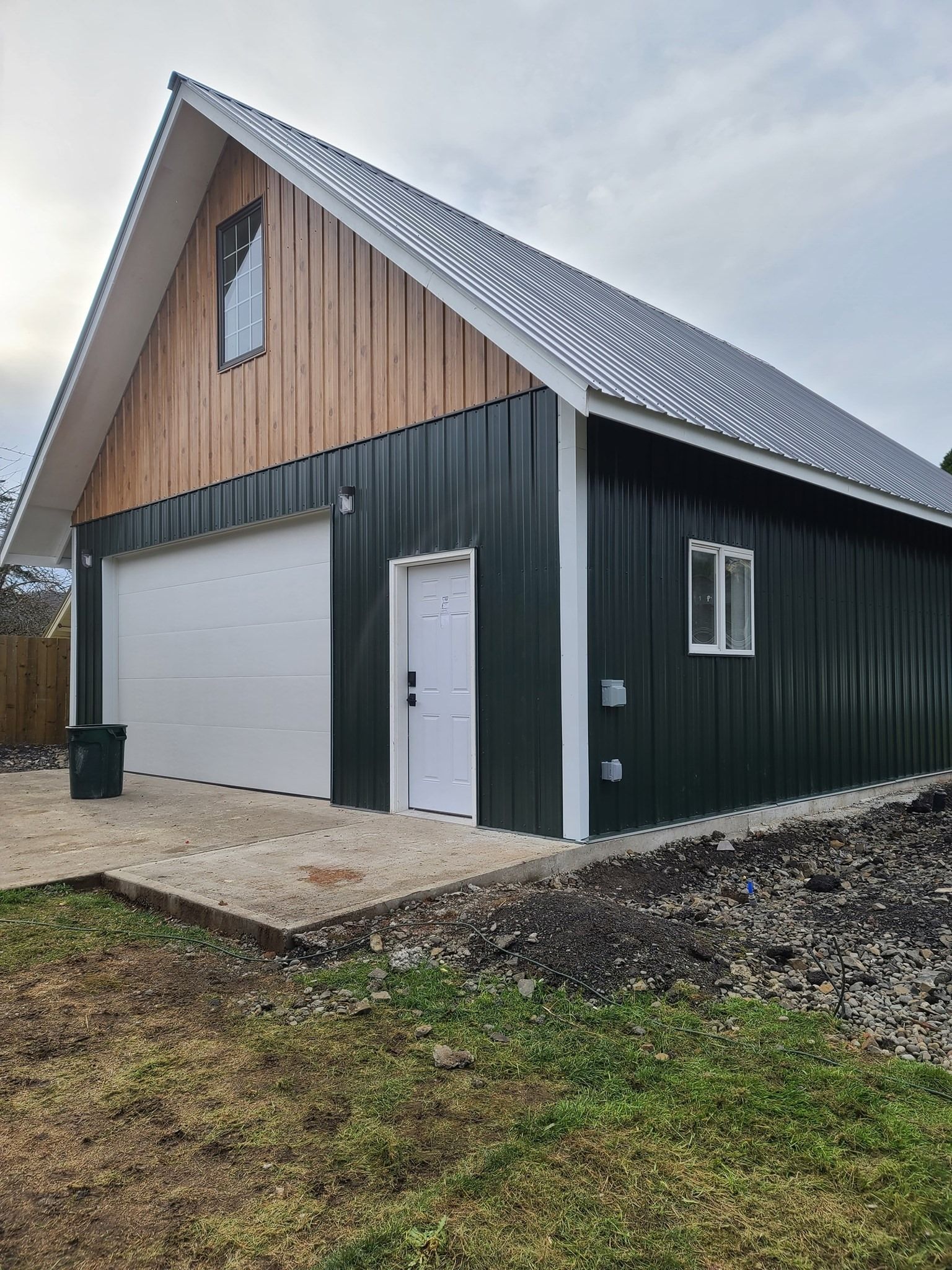 Green metal building with white garage door and wood accent under a cloudy sky.
